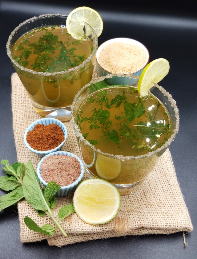 Masala Shikanji Recipe (Indian lemonade) in a whisky glass on jute mat with mint leave, lemon, sugar, salt and roasted cumin powder.