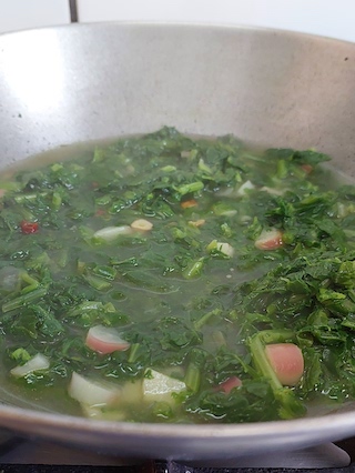 A stainless steel pot filled with colourful vegetables simmering on a gas stove burner. The vegetables include radish cubes. Steam is rising from the pot.
