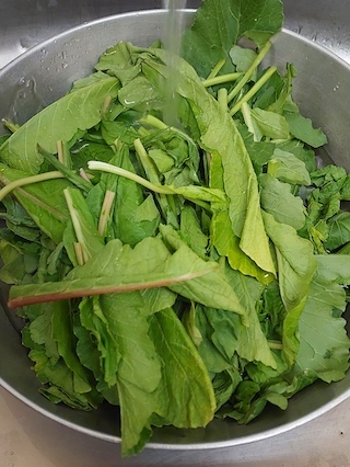 A colander filled with a variety of green leafy vegetables including spinach, kale, arugula, and swiss chard. The vegetables are being rinsed with water.
