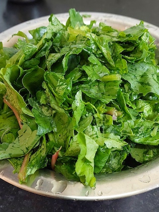 A stainless steel bowl filled with fresh spinach leaves. Some of the leaves have water droplets on them and there is water in the bottom of the bowl.
