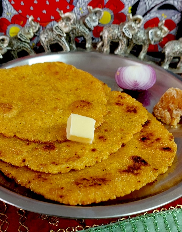 Image of freshly cooked Makki Ki Roti on a hot griddle, with golden-brown spots, served with a side of jaggery and onion piece.