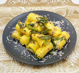 A plate of neatly rolled Khandvi garnished with fresh coconut, coriander leaves, and tempered mustard and sesame seeds.