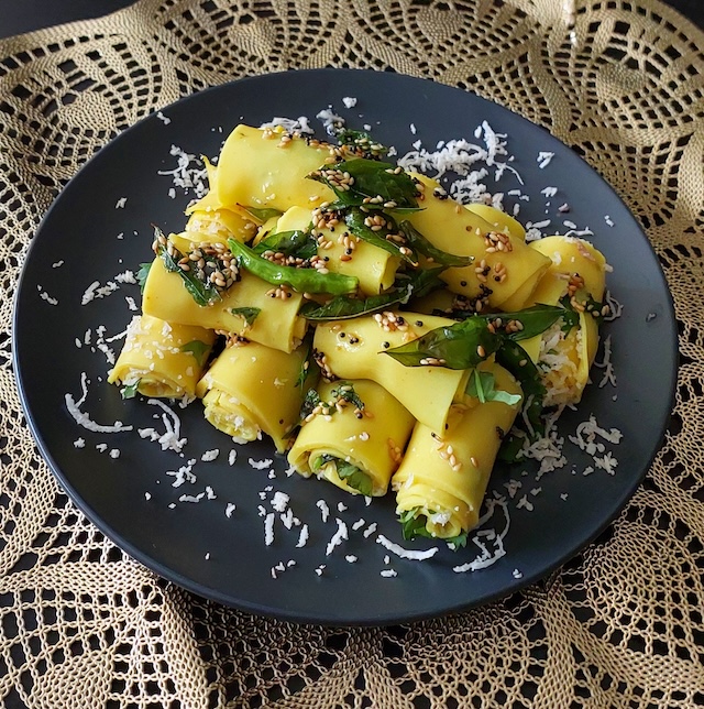 A plate of neatly rolled Khandvi garnished with fresh coconut, coriander leaves, and tempered mustard and sesame seeds.